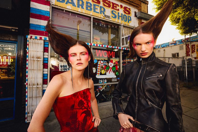 Two adult women with stylized makeup and hair pose outside a barber shop with colorful signage and red, white, and blue details.