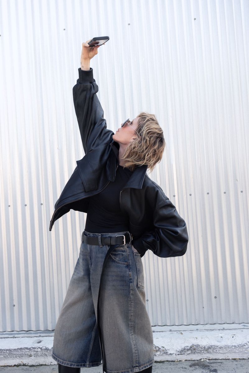 Woman in black jacket and culottes poses in front of corrugated wall, holding up her smartphone to take a selfie in daylight.