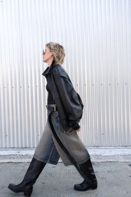 Stylish woman walks outdoors wearing black leather jacket, sunglasses, black top, wide denim shorts, black belt and black leather boots.