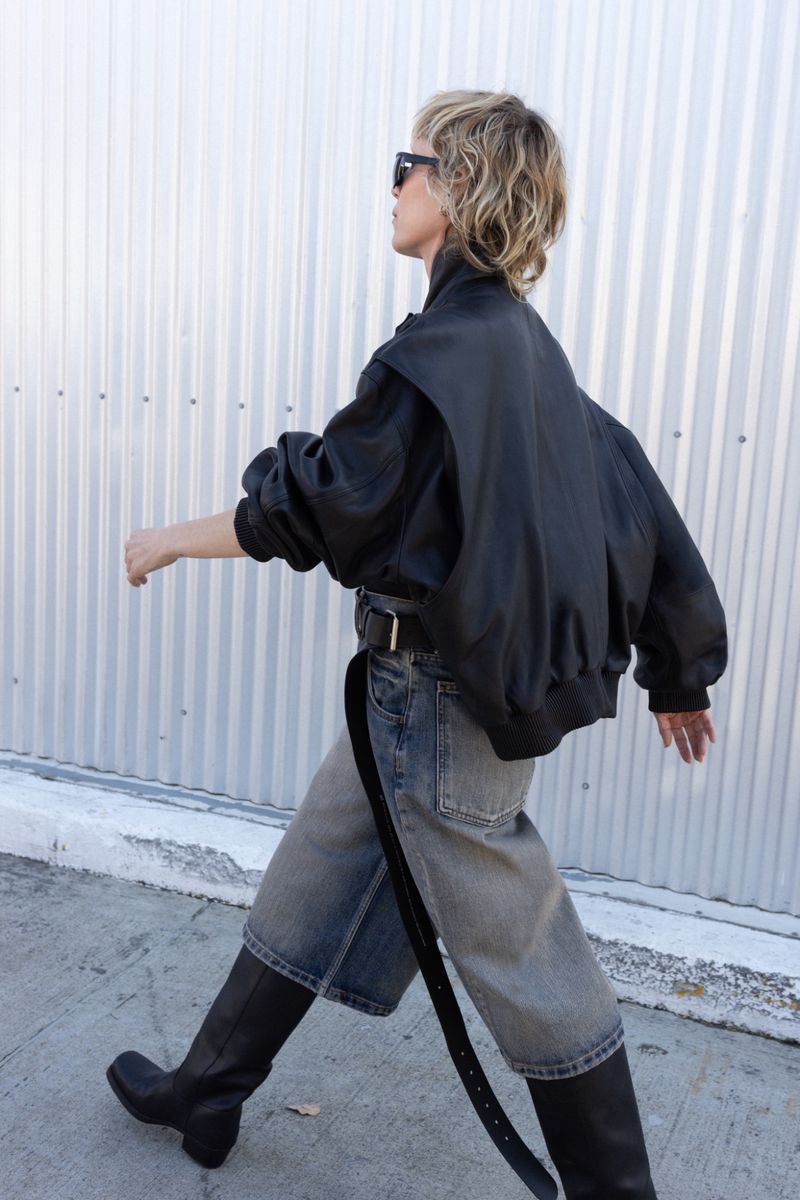 Adult female wearing a black leather jacket, denim shorts, and black boots walking against a white wall during the daytime.