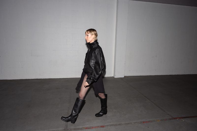 A slender woman in black leather jacket, dark skirt, fishnets and boots walks to the left in a stark urban setting with blank wall.
