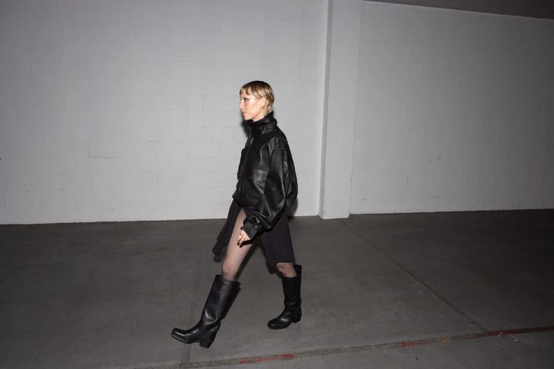 A slender woman in black leather jacket, dark skirt, fishnets and boots walks to the left in a stark urban setting with blank wall.