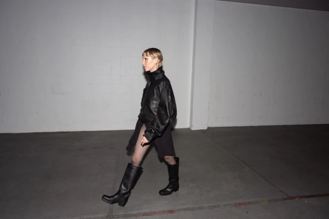 A slender woman in black leather jacket, dark skirt, fishnets and boots walks to the left in a stark urban setting with blank wall.