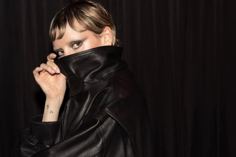 Close-up of a stylish adult woman with short blonde hair partially concealing her face with the high collar of a black leather jacket.