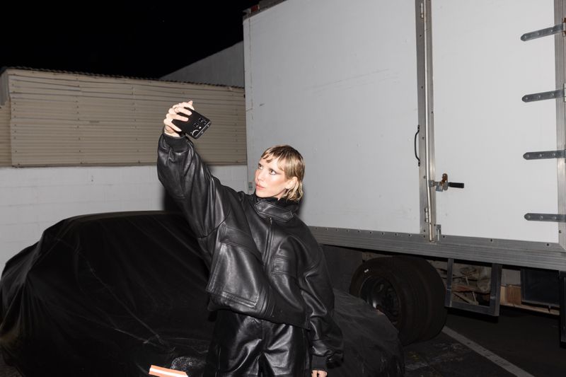 Woman in black leather outfit takes selfie outside near a covered car and a semi-truck in an urban environment at night.