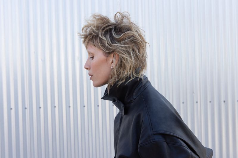 Profile of a blonde woman wearing a black leather jacket against a background of corrugated metal siding in an outdoor setting.