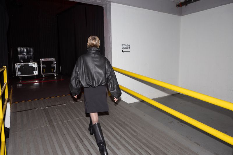 Adult female in a black leather jacket and boots walks in a backstage hallway with a stage door sign and black stage curtains.