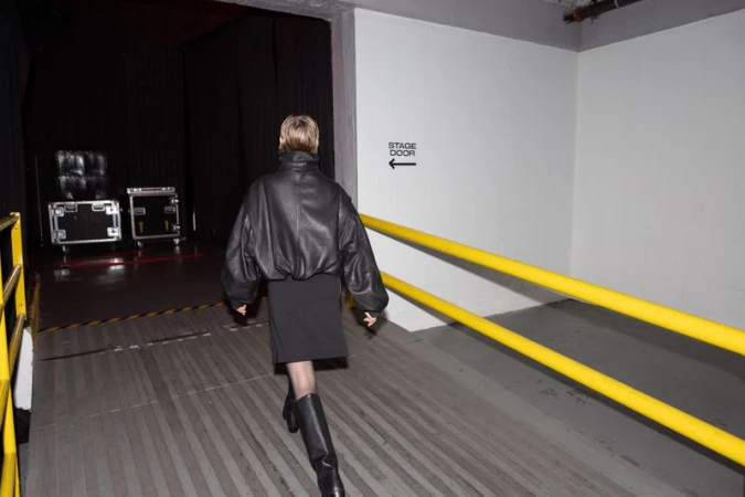 Adult female in a black leather jacket and boots walks in a backstage hallway with a stage door sign and black stage curtains.