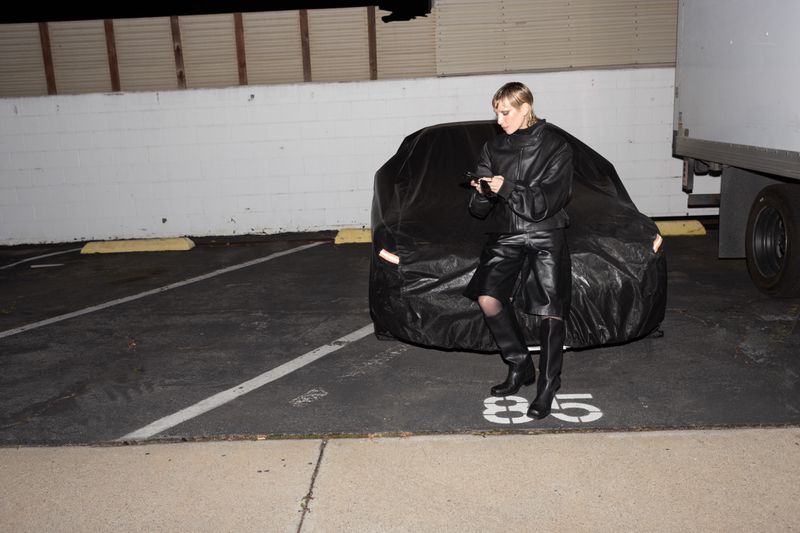 Woman in a black leather outfit poses next to a car covered in a tarp in an outdoor parking lot at night, using a cell phone.