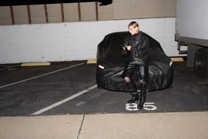 Woman in a black leather outfit poses next to a car covered in a tarp in an outdoor parking lot at night, using a cell phone.