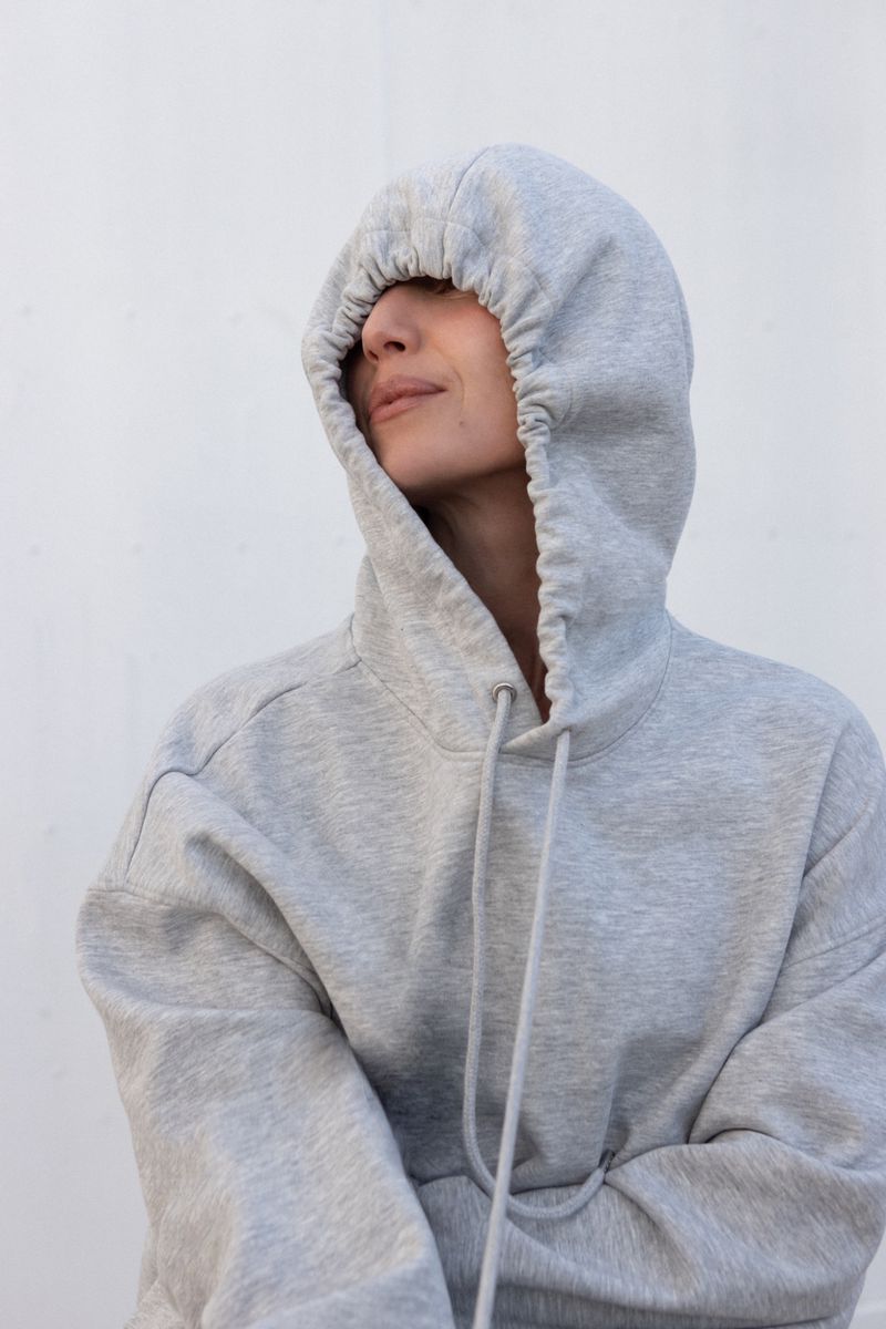 Woman wearing a grey hooded sweatshirt with the hood pulled over her head, posing against a plain white background.