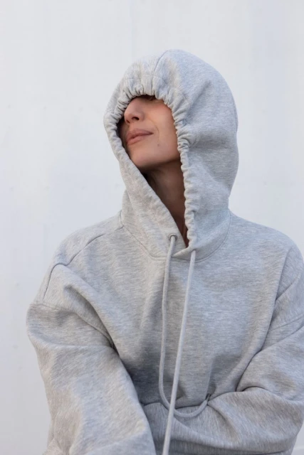 Woman wearing a grey hooded sweatshirt with the hood pulled over her head, posing against a plain white background.