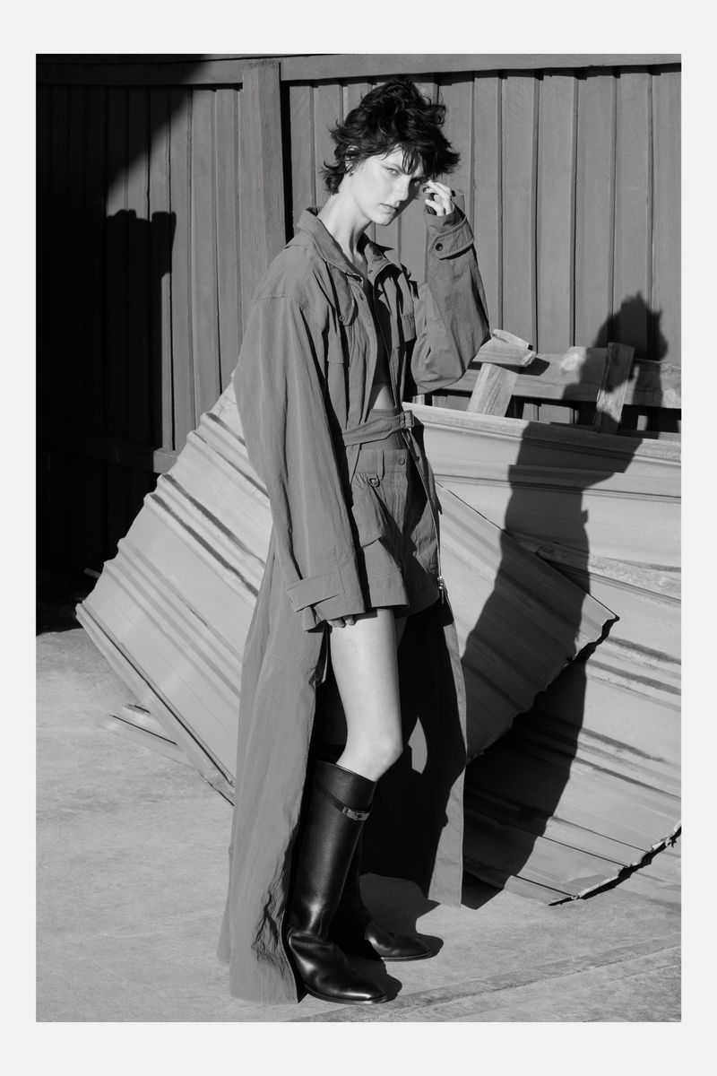 A woman in an oversized jacket and boots poses against a weathered metal wall, creating a high-contrast black-and-white editorial image.
