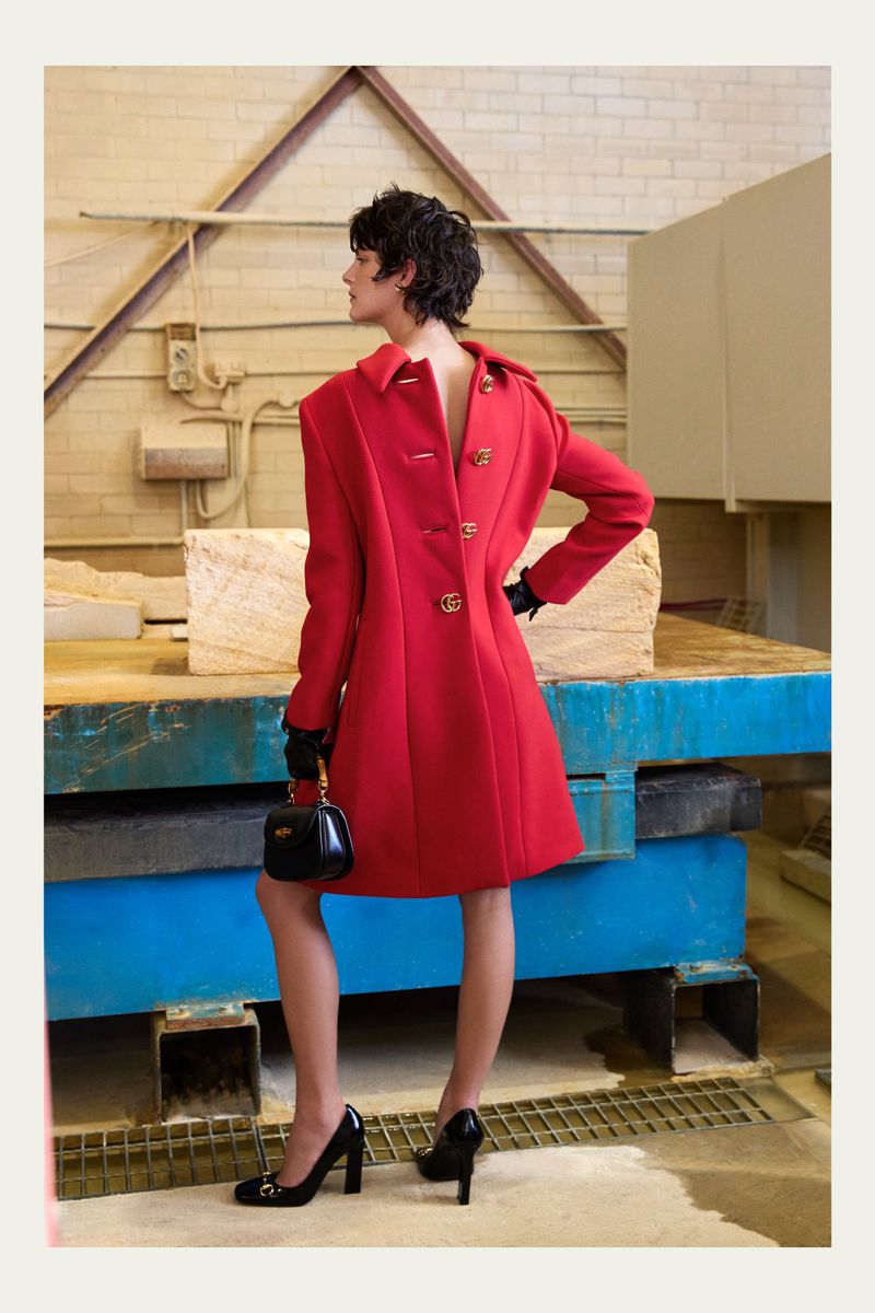 Brunette woman in red coat, black gloves, black heels and purse, poses indoors with stone objects and a white brick wall.