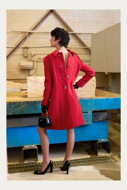 Brunette woman in red coat, black gloves, black heels and purse, poses indoors with stone objects and a white brick wall.