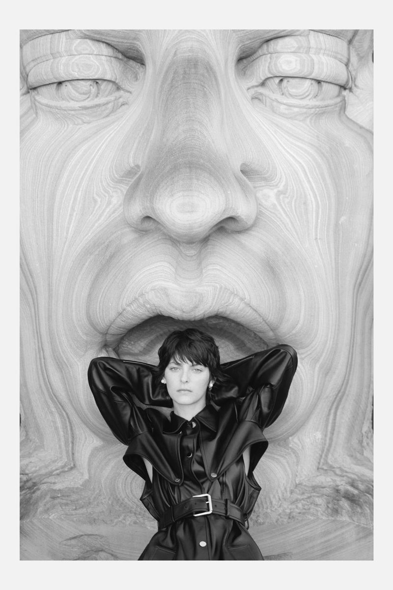 A woman in a black leather outfit poses in front of a large, monochrome wood carved face in an indoor setting with studio lighting.