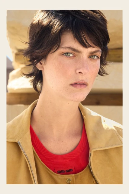 Close-up portrait of a young adult woman with dark hair and green eyes wearing a red shirt and tan jacket in natural light.