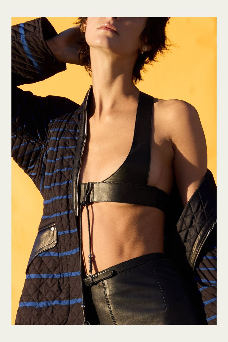 Fashion shot of a woman wearing a leather bra top and pants with a quilted jacket in front of a yellow background, lit by sunlight.
