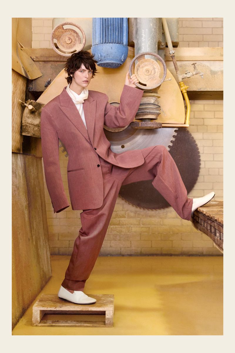 Adult fashion model poses in a mauve suit and white shoes against a backdrop of aged industrial machinery indoors with a yellow floor.