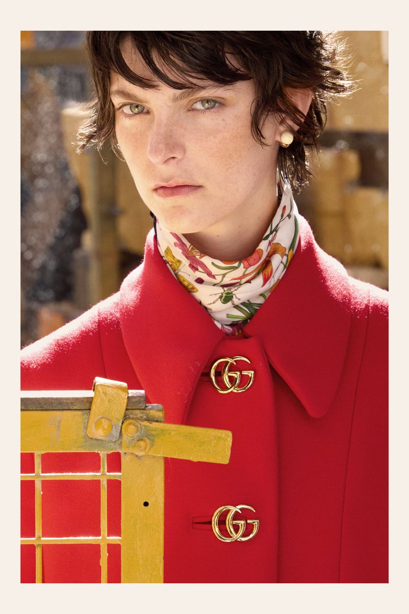Close-up shot of a model wearing a vibrant red Gucci coat with gold logo buttons, complemented by a floral scarf and pearl earrings.