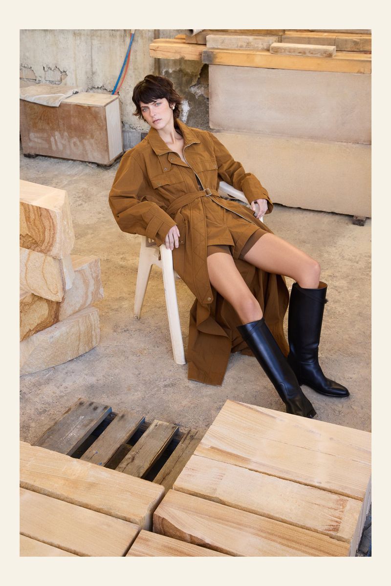 Fashion shot of a brunette woman in a brown trench coat and black boots sitting on a white chair in a rustic indoor location.