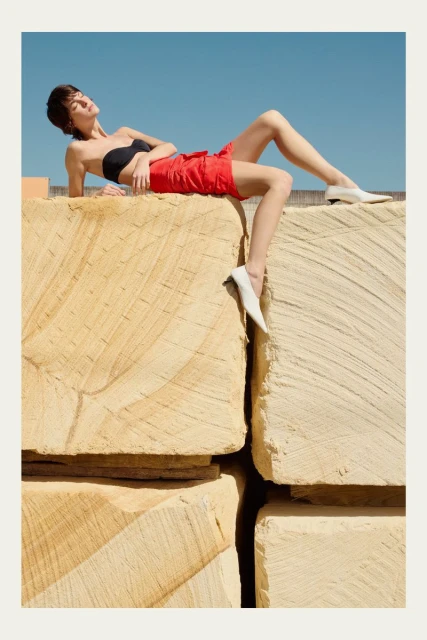 Woman in black tube top and red shorts sunbathing on stone blocks against a blue sky in an outdoor setting. Fashion photography.