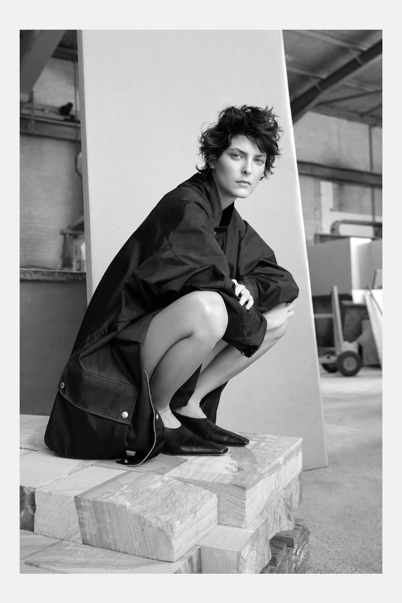 Black and white portrait of a woman crouching on stone blocks, wearing a dark coat and shoes, in an industrial setting.