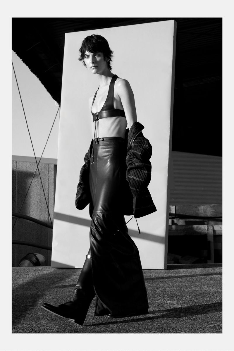 A woman in a dark outfit and leather bra and skirt is walking on a gray surface near a large white backdrop in a black and white fashion shot.