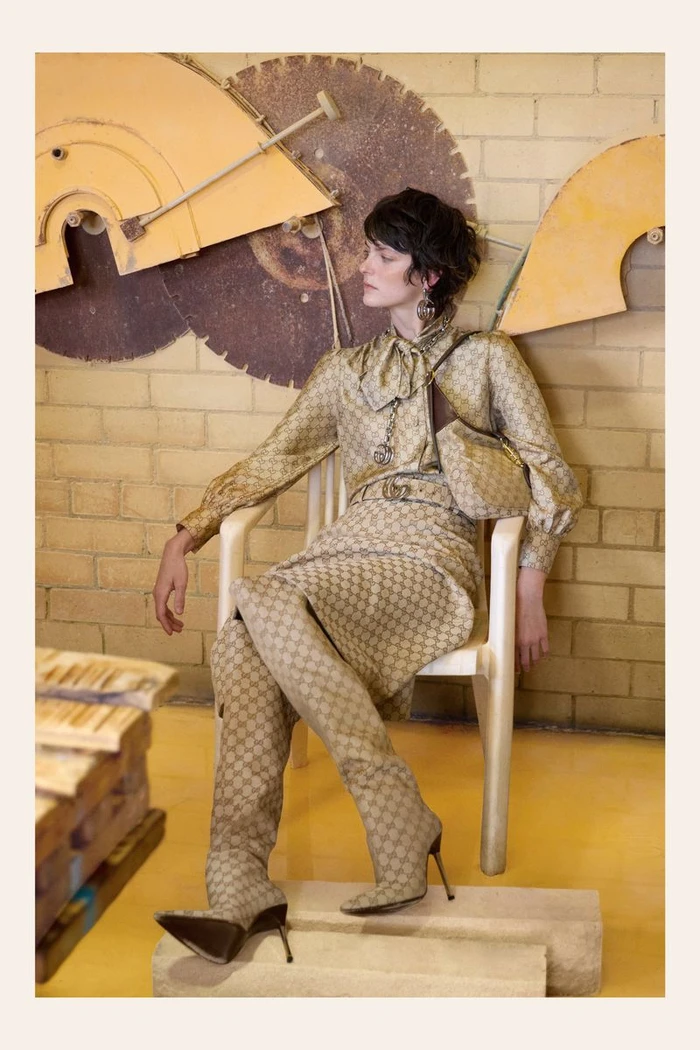 Model wearing a beige monogram print outfit and matching over-the-knee stiletto boots seated in an armchair against a yellow wall.