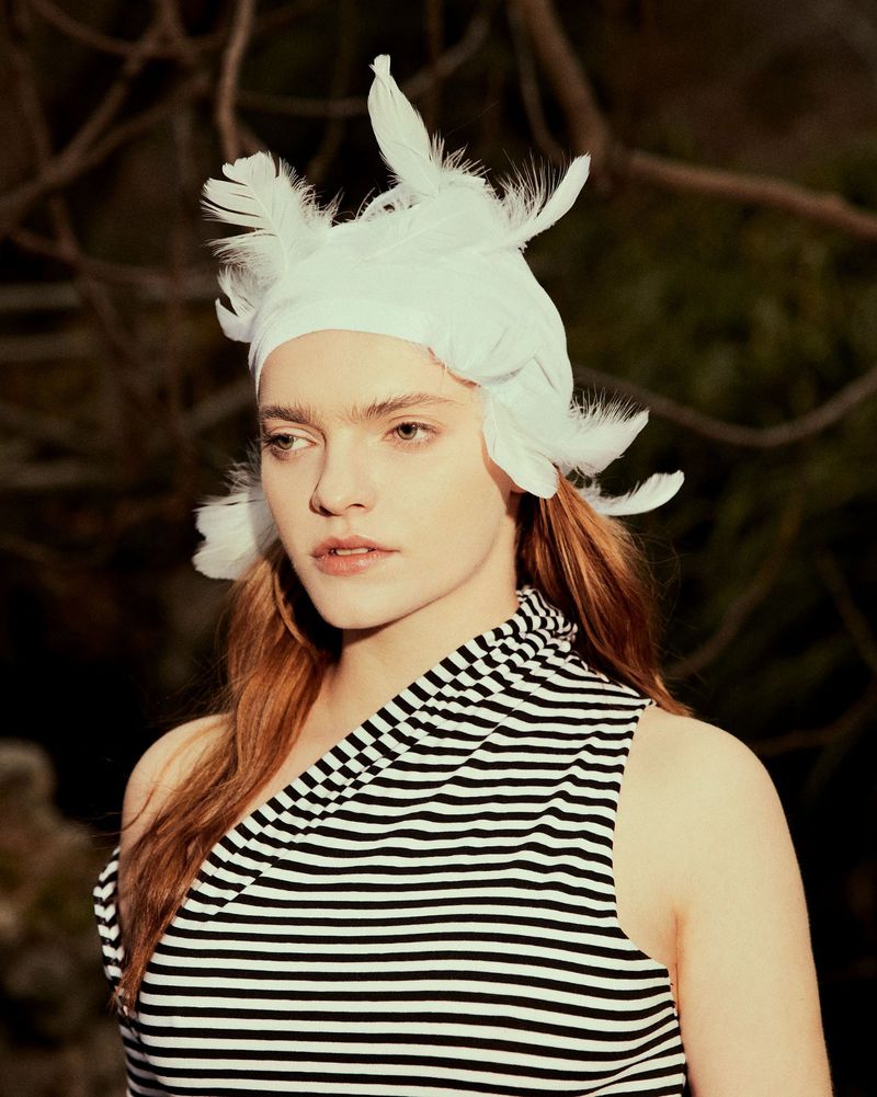 Portrait of a woman with red hair wearing a striped dress and a white hat adorned with feathers, set against a blurred outdoor backdrop.