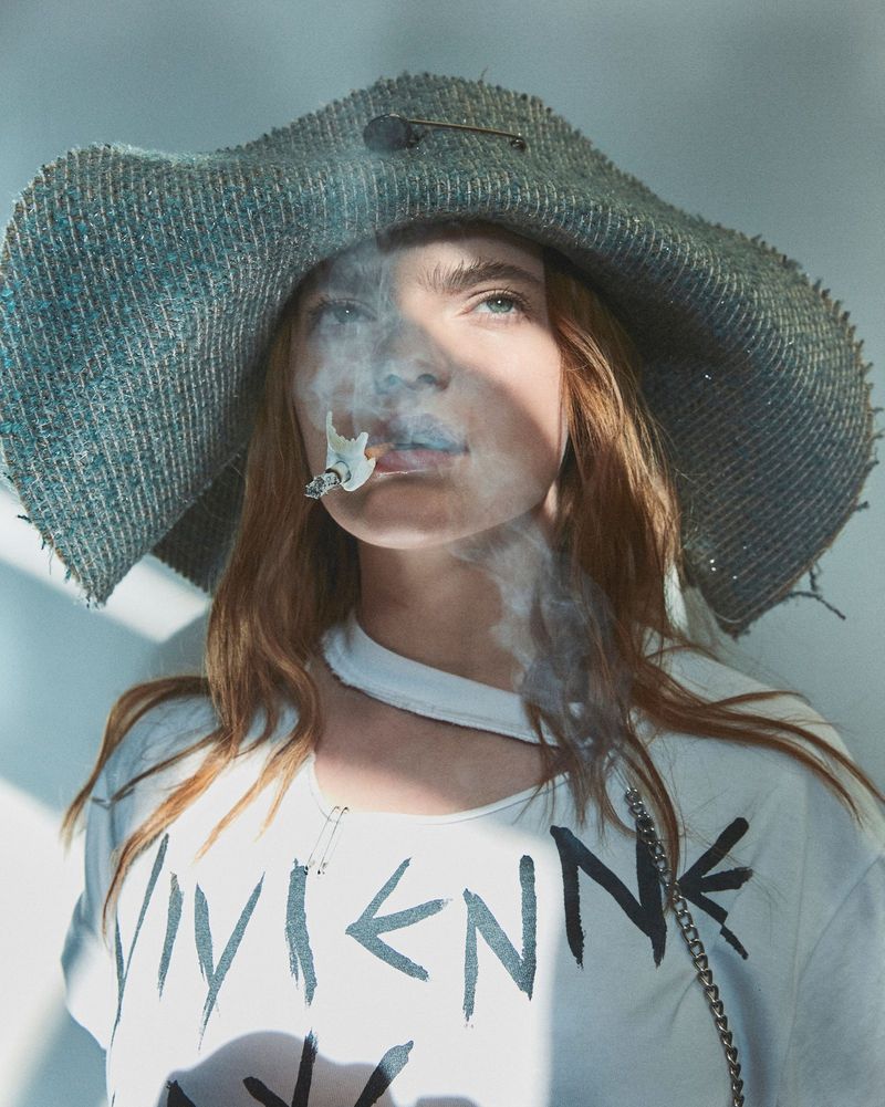 Stylish portrait of a woman with red hair wearing a hat and t-shirt, exhaling cigarette smoke against a neutral background.