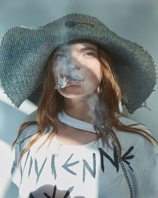 Stylish portrait of a woman with red hair wearing a hat and t-shirt, exhaling cigarette smoke against a neutral background.