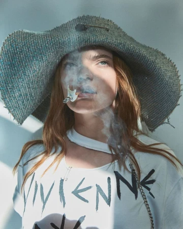 Stylish portrait of a woman with red hair wearing a hat and t-shirt, exhaling cigarette smoke against a neutral background.