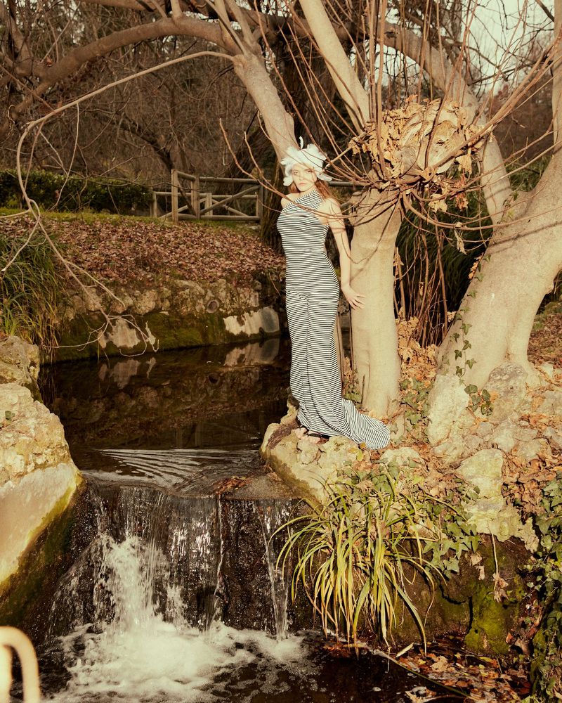 A woman in a striped dress poses near a small waterfall in a natural outdoor setting with trees and lush foliage, captured at eye-level.