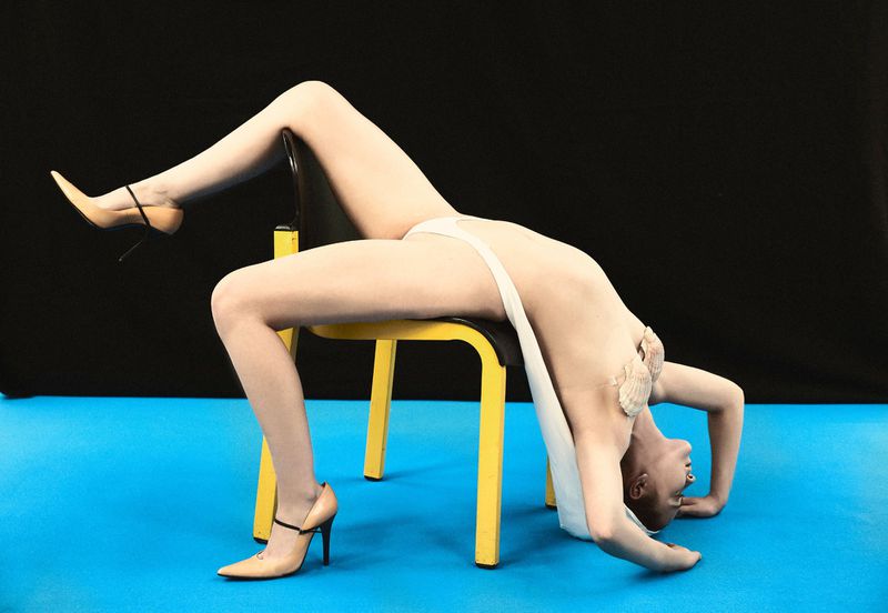 Adult female in artistic pose bridging over yellow chair in beige high heels against blue and black studio backdrop.