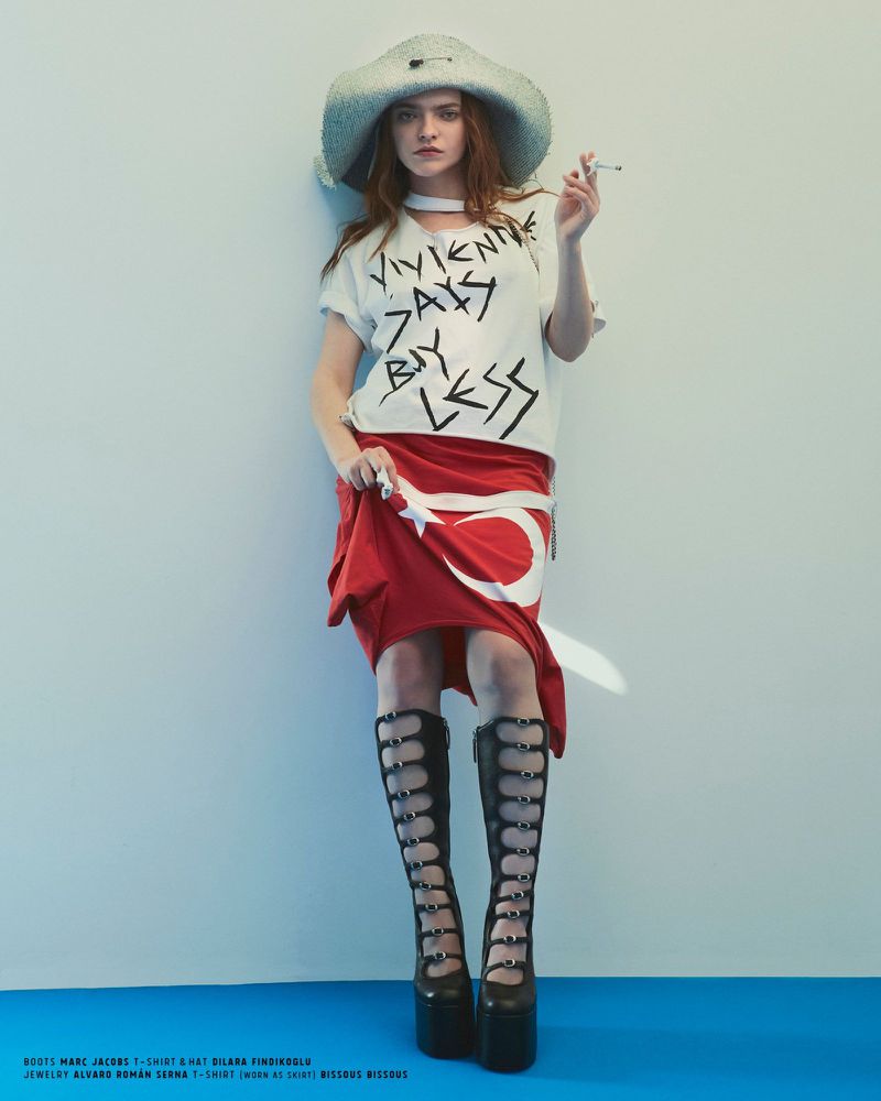 A woman poses in avant-garde fashion with a hat, graphic tee, Turkish flag skirt, and platform boots against a blue background.