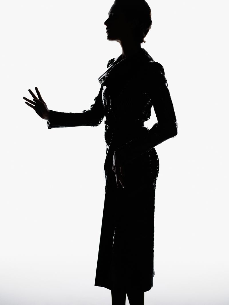 Silhouette of a woman in a dark coat and long pants against a white background with her arm outstretched and fingers spread.