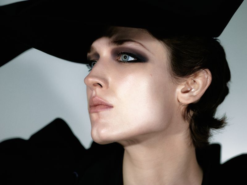 Portrait of a woman with dramatic smokey eyeshadow, wearing a large black hat. Shot in a photo studio with soft lighting and a white background.