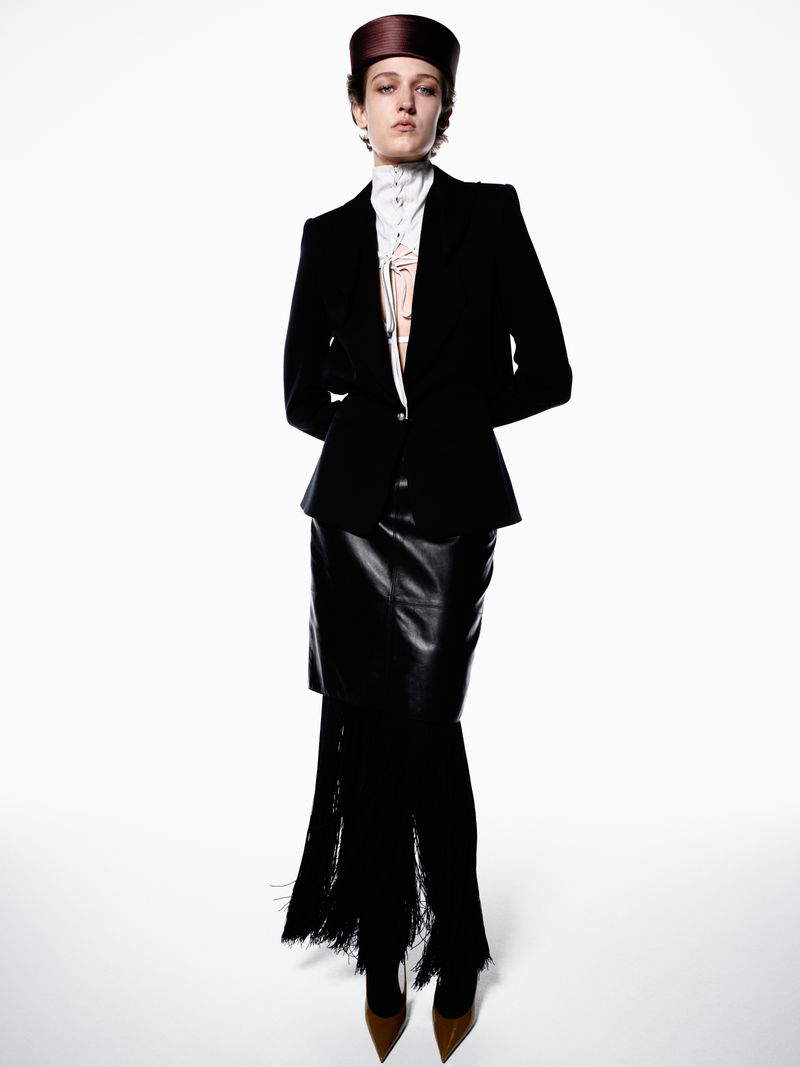 Full-length studio fashion shot of a woman in a black suit and leather skirt with fringe, against a white backdrop, stylish and elegant.