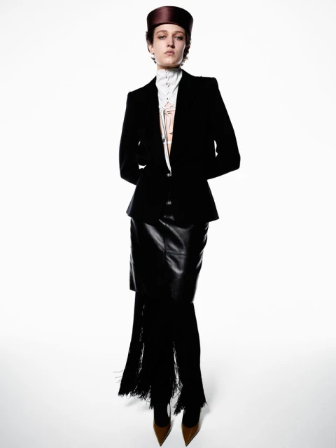 Full-length studio fashion shot of a woman in a black suit and leather skirt with fringe, against a white backdrop, stylish and elegant.