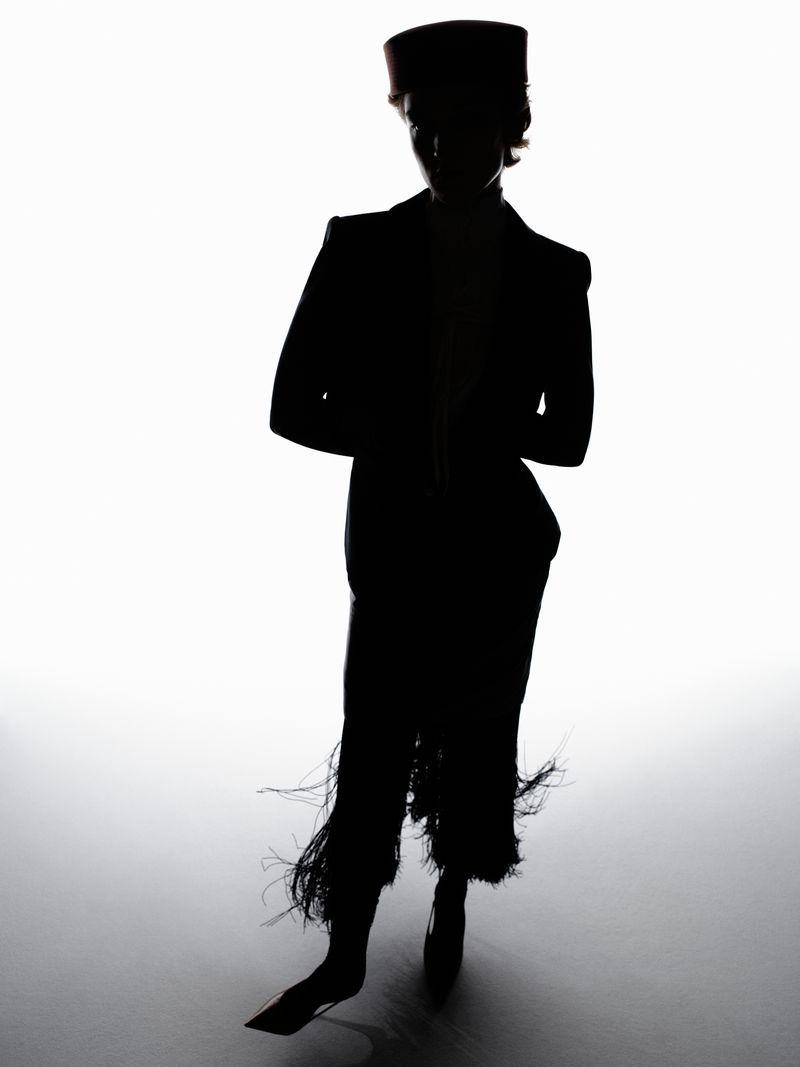 Silhouette of a person in a dark suit with fringed trousers and a hat against a bright white background in a high-contrast fashion shot.