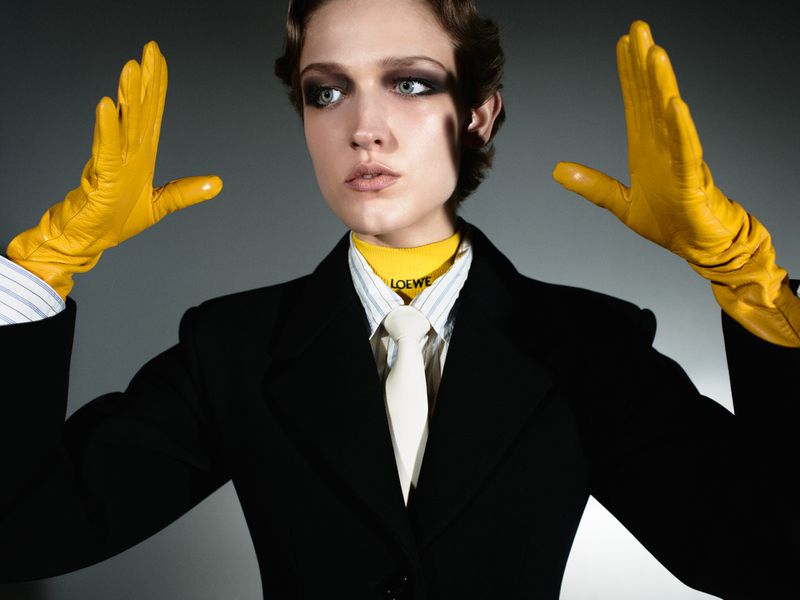 Stylish woman in black suit, yellow gloves, and necktie with LOEWE logo, posing in a studio setting, fashion photography, portrait