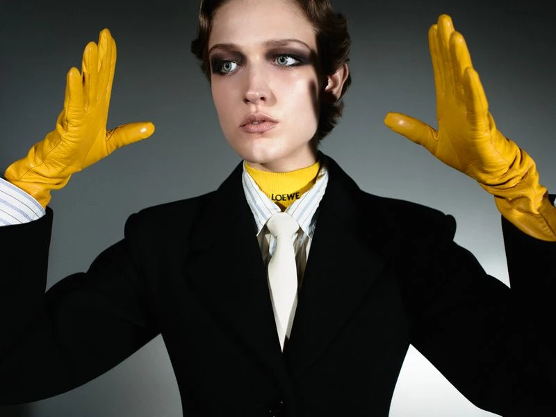Stylish woman in black suit, yellow gloves, and necktie with LOEWE logo, posing in a studio setting, fashion photography, portrait