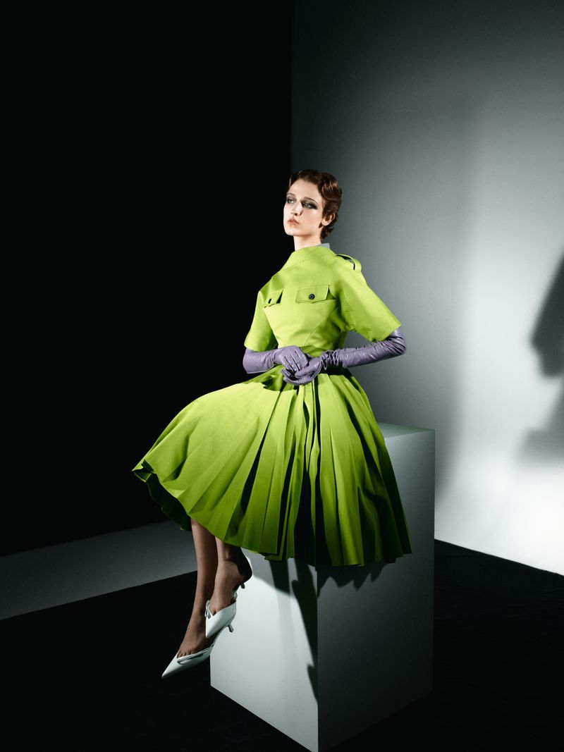 A young woman poses in a green dress and lilac gloves while seated on a white block against a two-tone gray and black background.