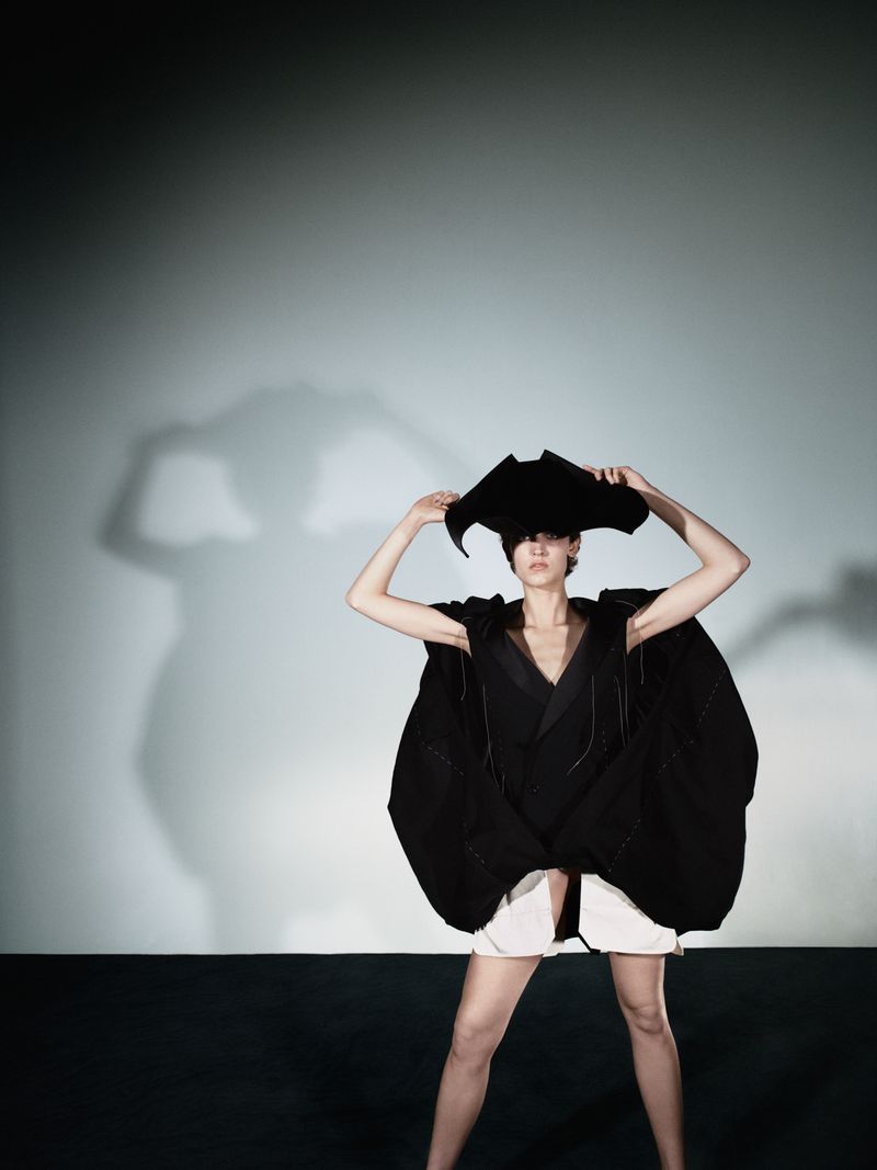 Fashion shot of a stylish woman in an avant-garde, dark, oversized dress and matching hat, posing in a studio with a clean backdrop.