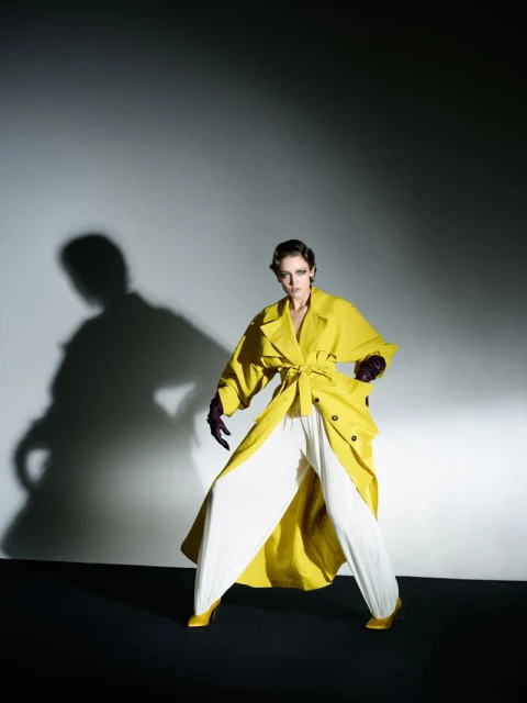 Woman in a yellow trench coat and white pants poses in front of a grey wall with a shadow. Fashion editorial style portrait.