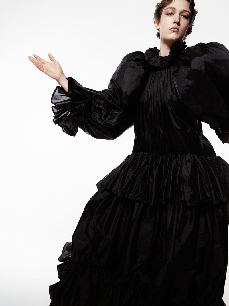 A woman wearing a large black ruffled dress with voluminous sleeves poses against a bright white backdrop in a fashion-style photograph.