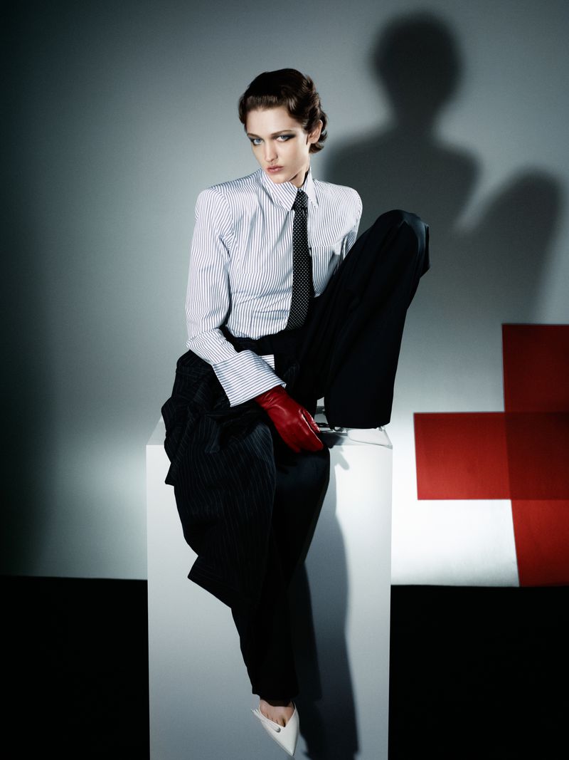 Fashionable woman in striped shirt and black trousers poses on white cube, wearing red gloves, captured in a stylish indoor studio setting.