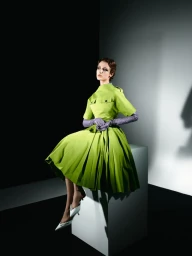 Model seated on a white block wearing a lime green dress and purple gloves against a split black and white studio backdrop