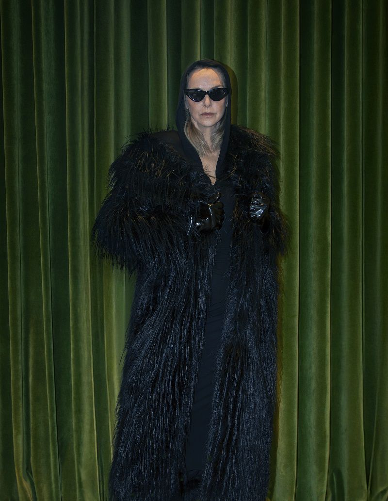 Elegant adult female in black fur coat and sunglasses posing against green curtain backdrop, high-fashion studio portrait. 